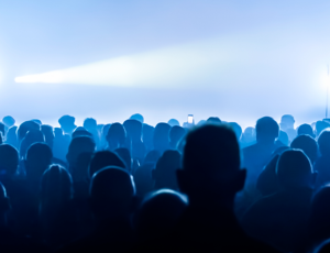 Crowd of people silhouetted in blue lighting at a concert or live event with bright stage lights in the background