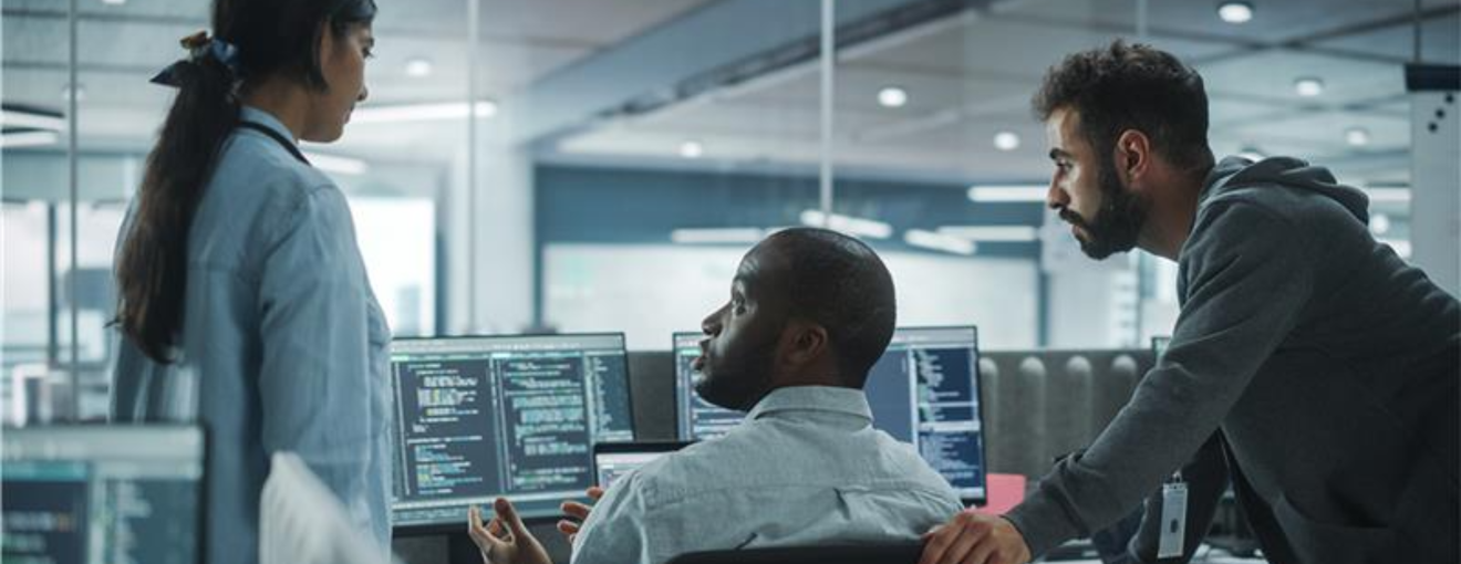 Team of IT professionals collaborating at multiple computer screens displaying code in a modern office